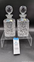 Front view of the matching pair of Bohemia crystal decanters with stoppers on a clear stand showing labels and intricate cut design.