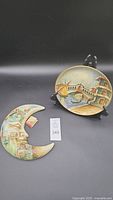 Two ceramic decorative items: a round plate with Venetian canal scene and a crescent moon with Venetian landmarks. Displayed against black background on stand.
