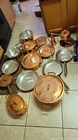 Overview of multiple copper-clad pans, saucepans, stock pot, fondue set and stainless interiors arranged on tile floor