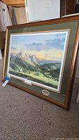 Angled view of the framed print leaning on carpet, showing wooden frame and scenic valley landscape.