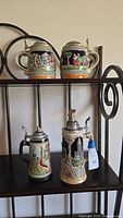 Four German hand-painted beer steins displayed on a shelf, showing varying sizes and detailed scenes on each stein