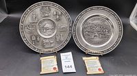 Two German pewter plates on stands with certificates laid in front, showing detailed embossed scenes and inscriptions on the plates.