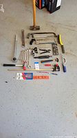 Full overview of assorted hand tools laid on garage floor including saws, wrenches, hammers, pliers, and measuring tools