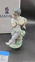 Porcelain figurine of boy with bongo drum, showing side view and details of figurine and original box in background.