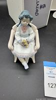 Front view of the Lladró figurine depicting a seated girl holding a small bouquet in a chair, original box in background