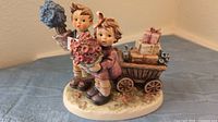 Front side of the M.I. Hummel figurine showing two children, one holding flowers and a letter, the other holding a flower pot, with a wagon behind them carrying gifts.