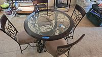 Full view of round glass top table surrounded by four matching chairs showing scrollwork metal frames and beige upholstered seats