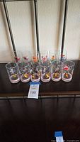 Set of ten glass tumblers and cups arranged on brown surface with whimsical animal and character paintings on each glass.