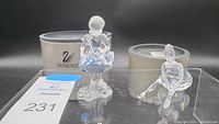 Photo showing two Swarovski ballerina figurines side by side on a black surface with original packaging in background.