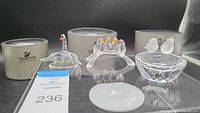 Three Swarovski crystal bird figurines displayed with their original cylindrical grey packaging in the background. Pieces show a swan, four small birds on a branch, and two birds on a bowl-shaped base.