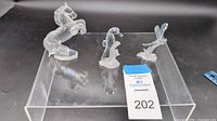 Top view of three clear Swarovski crystal figurines set: stallion, seahorse, dragonfly on a clear display stand with auction lot tag 202.
