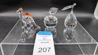 Three Swarovski crystal figurines displayed on clear acrylic stand: dinosaur with orange plates, faceted apple with leaf, faceted pear with leaf.