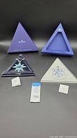 Two Swarovski triangular presentation boxes with a clear crystal star and snowflake ornaments displayed, including two small informational booklets and an auction card.