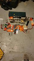 Photo showing assortment of tools, hardware packages, green plastic hardware organizer, drill bits, work light, and orange extension cord arranged on concrete floor.