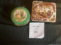 Two vintage trinket boxes side by side: one round with a courting couple painted on a green background lid, the other rectangular with colorful birds and floral design.