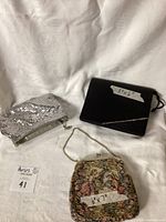 Photo showing three vintage clutches: silver bag, black clutch, and tapestry clutch on white fabric.