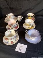 Seven fine bone china cups and saucers displayed on black cloth, showing floral patterns and gold trim.