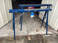 Two folding saw horses standing open on concrete beside a metal building door. Blue metal with black grips on top, showing front and side profiles.