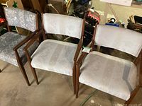 Three Mid-Century Modern dining chairs showing different upholstery styles and wooden frames, placed on a concrete floor with some surrounding clutter visible.