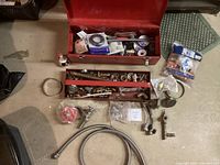 Photo showing open red metal toolbox with assorted plumbing accessories including rolls, fittings, and tools, with items spread out in front.