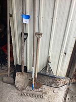 Garden tools standing against a shed showing five tools including shovels and rakes with visible surface rust