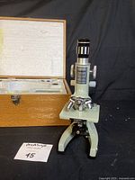 Front view of vintage Tasco microscope with dovetailed wooden carry case in background showing foam padding and accessories inside.