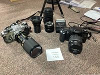 Photo showing the Canon AE-1 film camera with attached Leica telephoto lens, various lenses, Canon EOS camera with lens, and a flash unit arranged on carpeted floor.