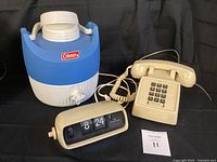 Photo of the 1970s blue and white Coleman jug alongside the Hamilton flip clock and beige touch tone desk phone on black background