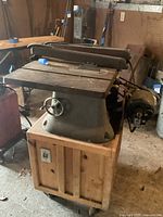 Front right angled view of beige metal Beaver Model 3200 table saw on wooden stand with casters, showing saw blade, fence, hand wheel, and motor attachment.