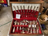 Wooden chest open showing flatware pieces arranged in velvet interior slots, including knives, forks, and spoons in multiple patterns.