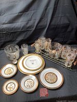 Photo showing a collection of vintage glassware and gold trimmed plates on a black cloth backdrop, including frosted glass bowl, clear gold leaf glasses in metal holder, and several decorative plates with gold leaf patterns.