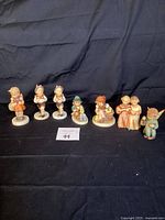 Photo showing all seven vintage Hummel figurines in a row on black fabric background for lot overview.