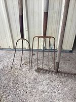 Two pitch forks and a garden rake leaning against a metal shed wall, showing handles and tines with surface rust.