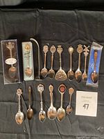 Full view of 13 silver collector spoons laid out on black background, showing variety of designs and some original packaging