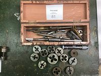 Open wooden toolbox with various metal threading tools inside and multiple round metal dies arranged on surface outside the box.