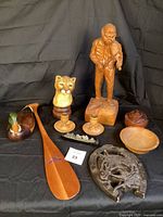 Photo of the entire lot showing wooden fiddler statue, wooden paddle, Mallard wood duck decoy, vintage cast iron railroad spike with mining locomotive, horseshoe wall decor, wooden carved lion head, wooden bowl, two candle holders, and lidded container arranged on black background.