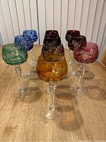 Top view showing 10 crystal wine glasses in various vibrant colors including cobalt blue, green, burnt orange and multiple shades of red and purple. Each glass features intricate white etching.