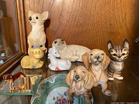 Collection of animal figurines displayed, including porcelain dachshunds, small ceramic cow and horse, stone resin seals, kitten and other animal figures.