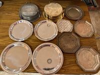 Overall view of the lot showing multiple silver and gold colored coasters stacked and some trays on a wood surface. Some tarnish visible on items.