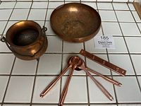 Small copper cauldron, pressed copper bowl, and measuring spoons with wall hanger displayed on white tiled surface.