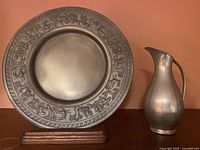 Decorative pewter plate with hunting party design on wooden stand next to Royal Guild pewter pitcher.