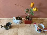 Four small decorative planters and vases arranged on wooden surface against a brown wall, including a white swan, a red slim vase with faux yellow roses, a painted ceramic watering can with floral design, and a brown glazed donkey and cart posy vase.