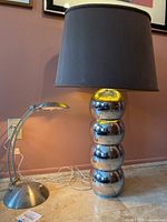 Two lamps side by side on a tiled floor against a pink wall showing the stacked chrome ball lamp with dark shade and the smaller silver adjustable lamp.