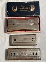 Top view of open original black and gold box with double harmonica inside and two additional harmonicas below on white background.