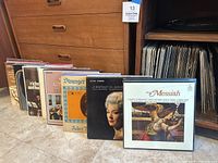 Front view of vinyl records displayed standing next to a cabinet, showing several album covers including Handel's Messiah and a portrait of a woman.