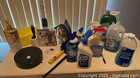 Various household cleaning products, detergents, a lint remover, and a black container seen on a table in front of closed blinds.