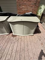 Image of closed heavy duty gray plastic outdoor storage container on wooden deck next to house exterior brick wall.