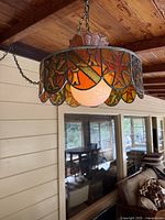 Side angled photo of hanging stained glass light fixture showing orange, red, and yellow floral mosaic glass panels and white center globe light diffuser under wooden ceiling