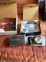 Panasonic Lumix DMC-ZS19 camera, carrying case, installation CD, and a $5 photo prints coupon all on a fabric surface.