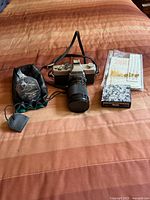 Full view of the Minolta SRT 200 camera set with accessories on orange-brown bedspread under natural light showing camera with lens, pouch, film box, case, and manual.
